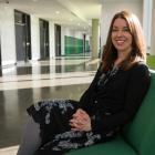 Smiling woman faces the camera, seated upon a green chair in a long corridor at an academic institution. She is dressed in a black blazer and patterned dress.