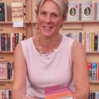 Woman smiles at the camera. She is in a pink top and wearing a necklace. In front of her is a pile of books, and in one hand is a pen.